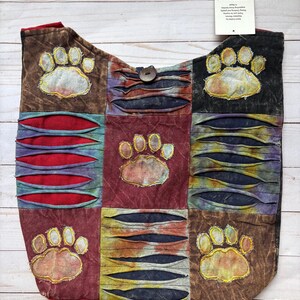 May include: A patchwork hobo bag featuring a multi-colored tie-dye design with five embroidered paw prints. The bag includes horizontal slashed fabric panels revealing red and dark blue layers, a single wooden button closure, and a white tag.