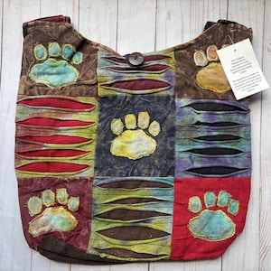 May include: A hobo bag featuring a patchwork design with brown, red, blue, and green fabric panels. The bag displays embroidered paw prints and horizontal cut-out details revealing contrasting fabric layers. A wooden button secures the top opening.