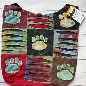May include: A patchwork shoulder bag featuring a multi-colored design with distressed horizontal slits and stitched paw print appliqués. The bag includes a wooden button closure and a white product tag with the text Rising and Made in Nepal.