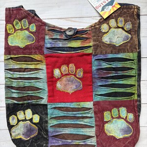 May include: A patchwork tote bag featuring a multi-colored tie-dye design with horizontal slashes and four embroidered paw print appliques. The bag includes a central button closure and a tag labeled Rising with the code RIB1727.