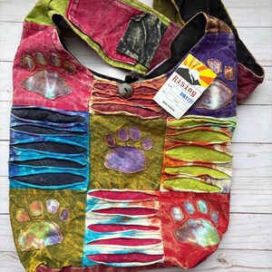May include: A colorful patchwork shoulder bag with paw print designs. The bag features a variety of colors including red, green, blue, and purple. It has a button closure and a tag that reads "Rising". The bag is made of a soft, textured fabric.