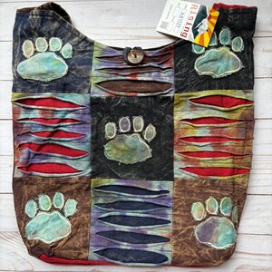 May include: A patchwork shoulder bag with a paw print design. The bag features squares of brown, black, and blue fabric with distressed accents and a button closure. The paw prints are in a light blue color.