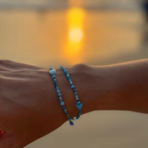 Pode incluir: Uma delicada pulseira de contas azuis com contas em forma de coração e redondas. A pulseira é uma mistura de tons de azul claro e escuro, criando um contraste subtil. É uma joia.