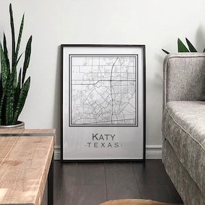 Könnte beinhalten: Ein schwarzer gerahmter Druck einer Karte von Katy, Texas, hängt an einer weißen Wand über einem grauen Sofa mit Kissen. Eine Topfpflanze mit Schlangenhaut steht auf einem Holztisch vor dem Sofa.