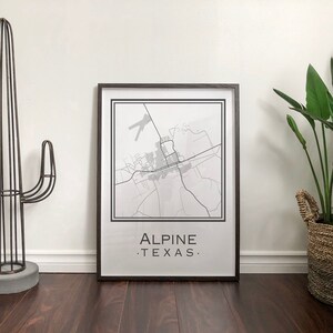 May include: A black framed print of a map of Alpine, Texas. The map is in black and white and shows the streets and roads of the city. The print is hanging on a white wall.