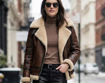 Puede incluir: Una chaqueta de cuero marrón con forro y cuello de borreguito color crema, sobre un jersey de cuello alto beige. El conjunto incluye pantalones negros ajustados con cinturón, botines negros y un pequeño bolso de mano negro.