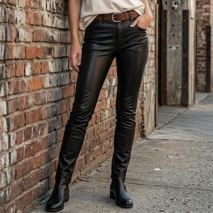 Puede incluir: Una camiseta de manga corta color crema combinada con pantalones negros ajustados y un cinturón de cuero marrón con hebilla plateada. El conjunto se completa con botines de cuero negro, creando un estilo coherente con tonos y texturas neutras.