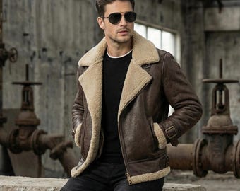Puede incluir: Una chaqueta de cuero marrón con un grueso forro de borrego beige, un cuello ancho con muescas y cierre de cremallera frontal. La chaqueta presenta mangas largas con puños de hebilla, un suéter negro debajo y pantalones oscuros con rodillas acanaladas.