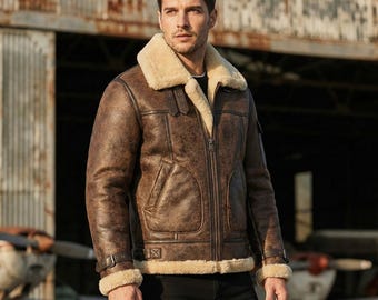 Puede incluir: Chaqueta de cuero marrón con cuello y puños de borrego. La chaqueta tiene cierre de cremallera, dos bolsillos delanteros y una hebilla en la cintura. El fondo muestra un hangar de aviones y aviones antiguos.
