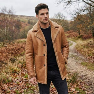 May include: A brown leather coat with a shearling collar and trim. The coat features a button closure and is worn over a black sweater and dark jeans. The background shows a path and trees, suggesting an outdoor setting.