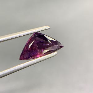 May include: A triangular-cut gemstone with deep purple and magenta hues, featuring multiple facets that reflect light. The stone is held by a pair of silver-toned metal tweezers, showcasing its translucent quality and sharp, geometric edges.
