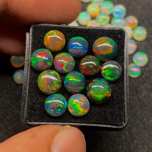 May include: A collection of round, polished opals in a black display case. The opals display a variety of colors, including green, blue, orange, and red, with a dark base color. Some opals are scattered outside the case.
