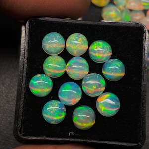 May include: A collection of round opals displayed in a black tray. The opals have a translucent, milky base color with vibrant flashes of green, blue, and orange, creating a play of color. The gemstones are polished and smooth, reflecting light.