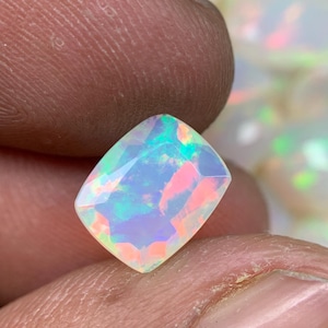 May include: A faceted cushion-cut opal gemstone displaying a translucent white base with vibrant flashes of neon pink, green, and blue play-of-color throughout its surface, held between two fingers for scale and clarity.