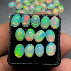 Op de afbeelding: Een verzameling ovaalvormige opalen, elk met een kleurenspel met iriserende flitsen van groen, rood en geel. De opalen zijn gerangschikt in een zwarte bak, met extra opalen zichtbaar op de achtergrond. Deze edelstenen zijn ideaal voor het maken van sieraden.