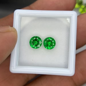 May include: Two round, brilliant-cut green gemstones displayed inside a small, square white plastic storage box. The stones feature a faceted surface that reflects light, showcasing a vibrant, saturated emerald green color and a circular shape.