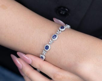 Op de afbeelding: Een zilveren armband met ovale, donkerblauwe edelstenen omringd door kleinere, heldere stenen. De armband wordt om een pols gedragen. De achtergrond is donker en effen.
