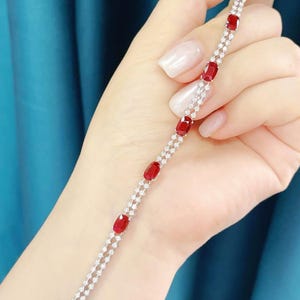 May include: A delicate bracelet featuring oval-cut red gemstones interspersed with small, sparkling white stones. The bracelet is set in a silver or white metal and is displayed against a blue background.