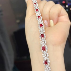 May include: A delicate bracelet featuring oval-cut red gemstones set in a silver-colored metal. The bracelet has a repeating pattern of red stones surrounded by smaller, clear stones, creating a sparkling effect. The design includes bow-shaped accents.
