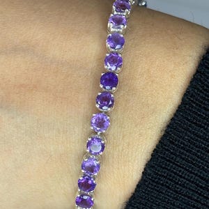 以下が含まれることがあります： 鮮やかな紫色の宝石が連なったシルバーブレスレット。宝石は互いに密接にセットされ、連続した色のラインを形成しています。ブレスレットは人の手首に装着されており、背景には黒い袖が見えます。