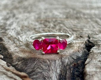 May include: A silver ring featuring a large, rectangular, bright pink gemstone, with two smaller heart-shaped pink gemstones either side. The ring band is detailed with small, clear stones.