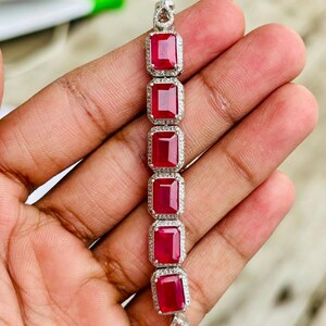 May include: A silver bracelet featuring six rectangular, vibrant red gemstones. Each ruby is framed by a halo of small, clear stones. The bracelet is held in a hand, showcasing its design and craftsmanship.