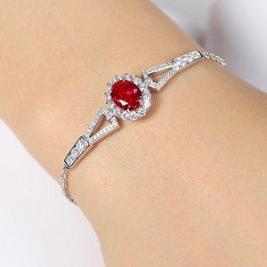 May include: A silver bracelet featuring a central oval-shaped, deep red gemstone surrounded by a halo of smaller, clear stones. The bracelet has a delicate chain and geometric accents with additional clear stones.