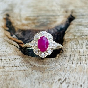 May include: A gold ring featuring a large oval ruby center stone, surrounded by a halo of smaller, round, clear gemstones. The ring's band has a delicate, textured design. The ring is set against a natural wood grain background.