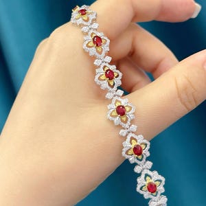 May include: A delicate bracelet featuring a series of floral-shaped links. Each link showcases a vibrant red gemstone at its center, surrounded by smaller, sparkling white stones. The bracelet's design incorporates gold and silver tones, creating an elegant and eye-catching accessory.