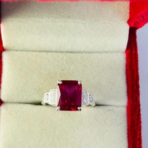 May include: A silver ring with a large, rectangular, ruby-red gemstone as the focal point. The ring is set in a red velvet jewelry box. The ring has smaller, clear gemstones on either side of the main stone.