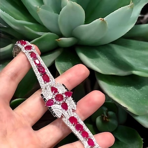 May include: A silver bracelet with a series of oval-cut red gemstones and smaller clear stones. The bracelet features a central design with a cluster of red gemstones and black accents. The background is a green succulent plant.