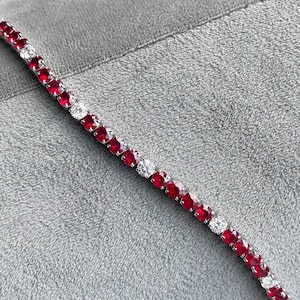 May include: A tennis bracelet featuring alternating red gemstones and clear, round-cut stones. The bracelet is set on a silver-colored metal and rests on a gray surface. The gemstones are evenly spaced along the bracelet.