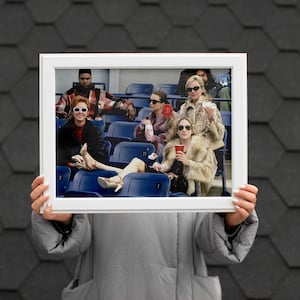 Op de afbeelding: Een wit omkaderde foto, vastgehouden door een persoon, met een groep mensen in een stadion. De mensen dragen zonnebrillen en jassen, sommigen houden drankjes vast. De achtergrond is een donker, getextureerd oppervlak.