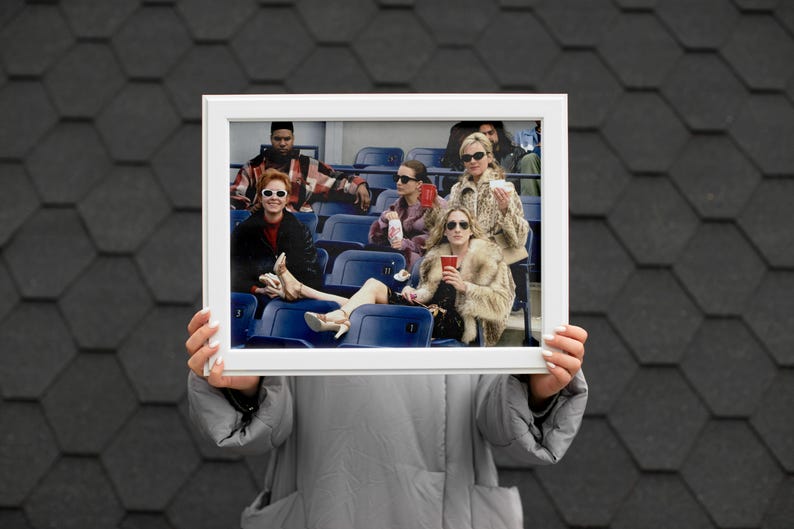 Op de afbeelding: Een wit omkaderde foto, vastgehouden door een persoon, met een groep mensen in een stadion. De mensen dragen zonnebrillen en jassen, sommigen houden drankjes vast. De achtergrond is een donker, getextureerd oppervlak.