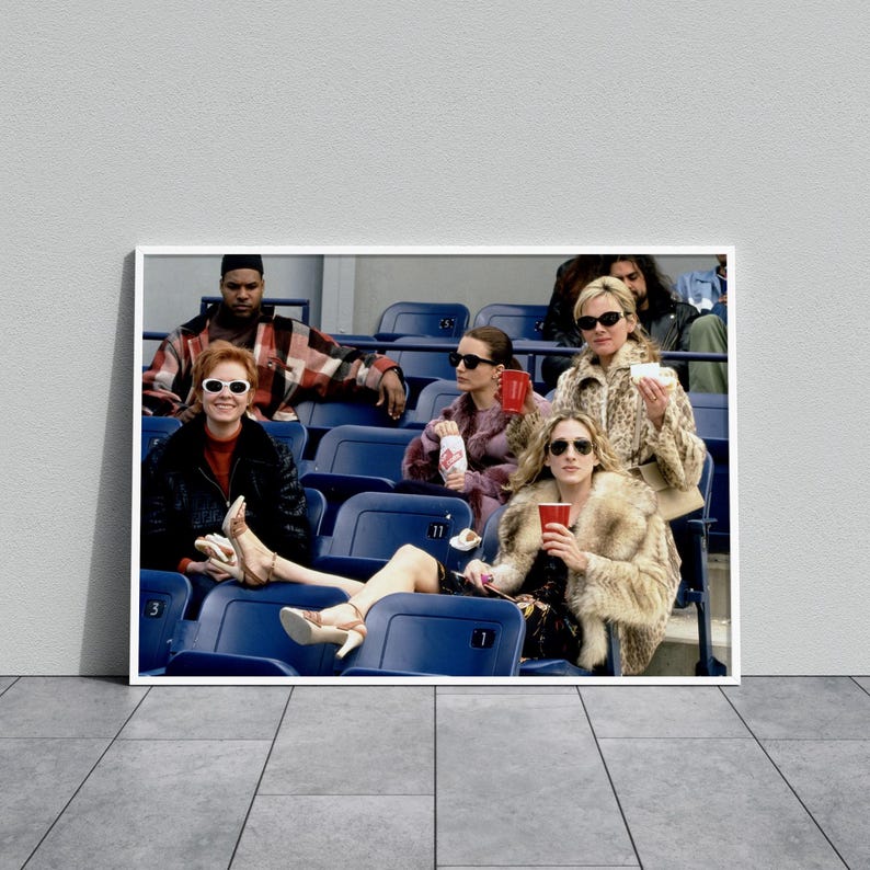 Op de afbeelding: Een ingelijste print met een groep mensen in een stadion. De personen dragen zonnebrillen en bontjassen en houden drankjes vast. De afbeelding is geplaatst tegen een grijze muur en vloer.