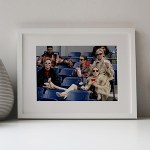 Op de afbeelding: Een ingelijste foto toont een groep mensen in een stadion. Ze dragen zonnebrillen en bontjassen en houden drankjes vast. De afbeelding staat tegen een witte achtergrond, met een vaas en een cactus op de voorgrond.