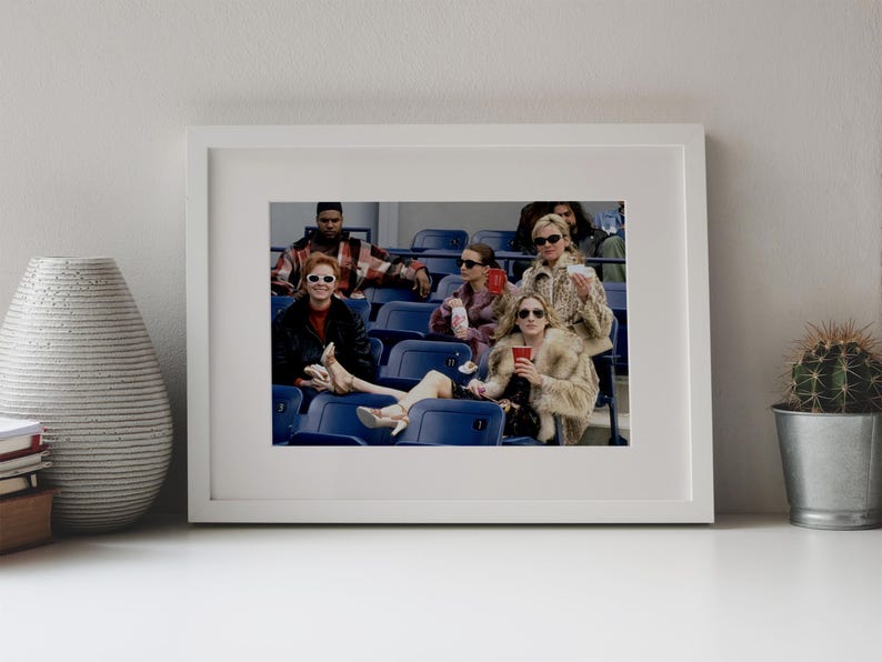 Op de afbeelding: Een ingelijste foto toont een groep mensen in een stadion. Ze dragen zonnebrillen en bontjassen en houden drankjes vast. De afbeelding staat tegen een witte achtergrond, met een vaas en een cactus op de voorgrond.