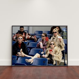 Op de afbeelding: Ingelijste print met een groep mensen in een stadion. De personen zitten in blauwe stadionstoelen, sommigen dragen zonnebrillen en bontjassen en houden drankjes vast. De afbeelding heeft een vintage, filmische uitstraling.