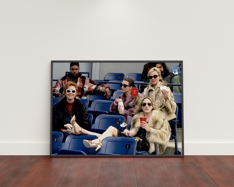 Op de afbeelding: Ingelijste print met een groep mensen in een stadion. De personen zitten in blauwe stadionstoelen, sommigen dragen zonnebrillen en bontjassen en houden drankjes vast. De afbeelding heeft een vintage, filmische uitstraling.