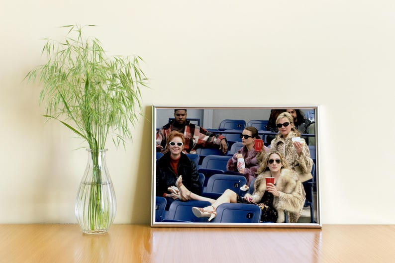 Op de afbeelding: Een ingelijste foto van een groep mensen met zonnebrillen en modieuze kleding, zittend in stadionstoelen. De afbeelding wordt weergegeven op een houten oppervlak, naast een glazen vaas met groen gebladerte.