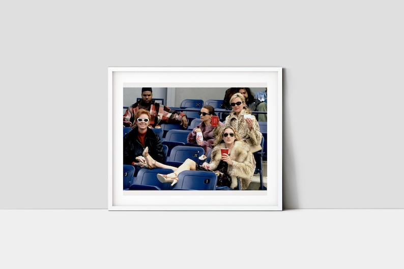 Op de afbeelding: Een ingelijste foto toont een groep mensen in een stadion. Verschillende personen dragen zonnebrillen en bontjassen, zittend op blauwe stadionstoelen. De afbeelding heeft een vintage esthetiek.