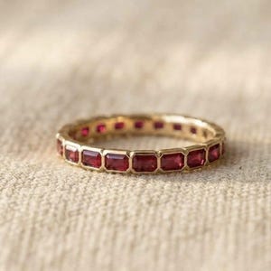 Könnte beinhalten: Ein Goldring als Eternity-Band mit einer durchgehenden Reihe tiefroter, im Smaragdschliff geschliffener Edelsteine in Zargenfassung. Der Ring zeigt eine gleichmäßige Anordnung rechteckiger Steine, die den gesamten Umfang des polierten Goldbandes umschließen.