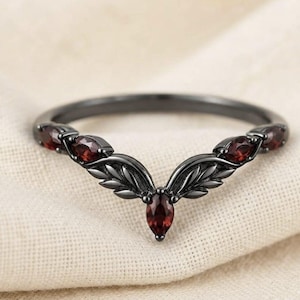 Peut inclure: Une bague en métal noir avec un anneau en forme de V au motif de feuilles. La bague est ornée de cinq pierres précieuses grenat rouge foncé taillées en marquise, dont une centrée à la pointe et deux plus petites de chaque côté de l'anneau.