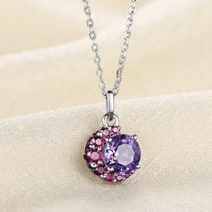 Peut inclure: Un collier argenté avec un pendentif orné d'une grande pierre précieuse ronde et facettée de couleur violette. Le pendentif est agrémenté d'un groupe en forme de croissant de petites pierres précieuses rondes rose foncé entourant la pierre violette.