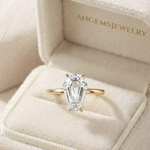 May include: A gold ring featuring a clear, coffin-shaped faceted gemstone set in a four-prong setting. The ring is displayed inside a cream-coloured velvet jewellery box with the text ANGEMSJEWELRY printed in gold on the inner lid lining.
