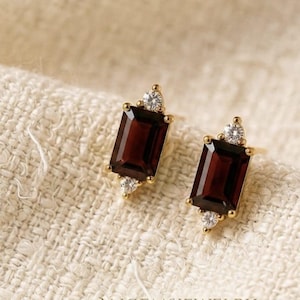 Könnte beinhalten: Ein Paar goldene Ohrstecker mit rechteckigen, im Smaragdschliff gehaltenen, dunkelroten Granat-Edelsteinen. Jeder Ohrring ist oben und unten mit zwei kleinen, runden, klaren weißen Diamanten verziert. Der Text ANGEMSJEWELRY ist unten sichtbar.