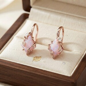 May include: A pair of rose gold drop earrings featuring a kite-shaped iridescent pink opal stone set within a delicate metal frame of sculpted leaves and vines, accented with a small clear gemstone at the top of each earring hook.