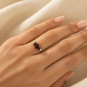 May include: A gold ring featuring a central hexagonal dark red gemstone flanked by two smaller clear triangular stones on each side. The band is thin and metallic, showcasing a refined design with a prominent deep red centre stone and clear accents.