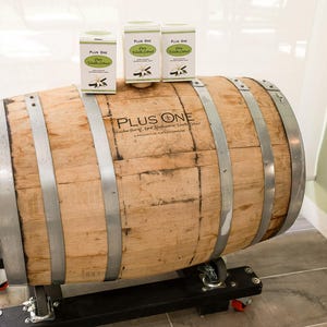 May include: A large wooden barrel with metal bands and the text "PLUS ONE" is displayed. Three boxes of "Pure Vanilla Extract" are on top of the barrel. The barrel is on a black metal stand with wheels.