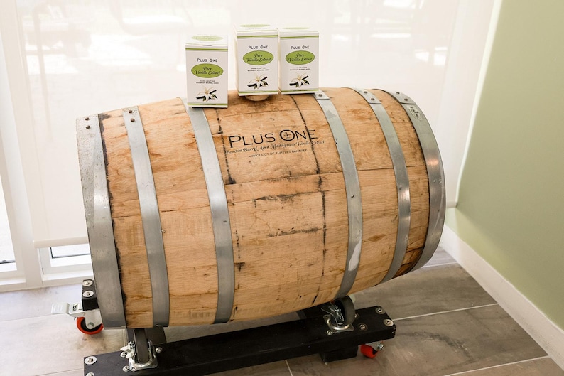 May include: A large wooden barrel with metal bands and the text "PLUS ONE" is displayed. Three boxes of "Pure Vanilla Extract" are on top of the barrel. The barrel is on a black metal stand with wheels.
